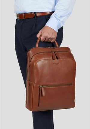 Mann i blå skjorte og mørke bukser som holder en brun lærmappe med frontlomme med glidelås og topphåndtak.