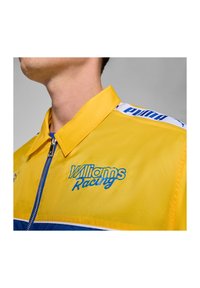 Zbliżenie na osobę w żółtej kurtce Williams Racing z niebieskim zamkiem błyskawicznym i logo Pumy na ramieniu.
