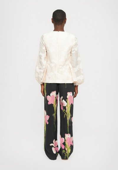 Lichtbeige top met textuur en pofmouwen, gecombineerd met zwarte broek met grote roze bloemenprints. Achteraan zichtbaar is de ritsdetails.
