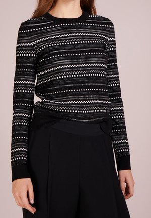 Frau trägt einen schwarzen Pullover mit langen Ärmeln, verziert mit weißen horizontalen gepunkteten und gestrichelten Streifen, kombiniert mit hoch taillierten schwarzen Hosen.