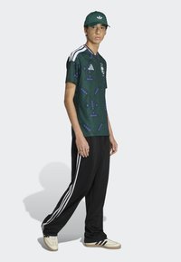 Green patterned athletic shirt with white and purple geometric details; black joggers with white stripes along the sides; beige sneakers.