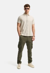 Beige kurzärmliges Text-shirt, olivgrüne Cargo-Hosen mit mehreren Taschen und beige Sneaker; Modell trägt schwarze Sonnenbrille.