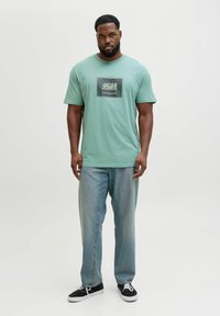 Miętowozielona koszulka z krótkim rękawem z ciemnym logo w formie graficznego prostokąta. W połączeniu z jasnoniebieskimi prostymi dżinsami i czarnymi trampkami.