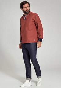 Camicia a maniche lunghe color ruggine con scollatura a V e taschino sul petto, abbinata a jeans in denim scuro e sneakers alte bianche. Visibili polsini a righe.