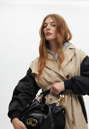 Una donna con lunghi capelli mossi indossa un trench beige e nero, mentre regge una borsa nera su uno sfondo bianco semplice.
