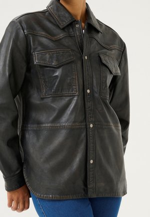 Person iført en sort læderjakke med frontknapper og to brystlommer med klap, kombineret med blå jeans.