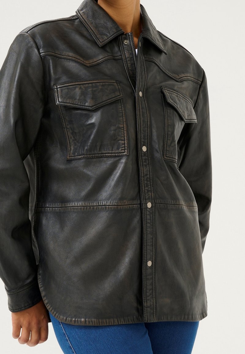 Personne portant une veste en cuir noire avec des boutons-pression à l'avant et deux poches à rabat sur la poitrine, associée à un jean bleu.