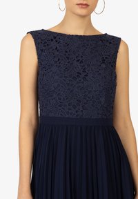Robe marine avec un corps en dentelle présentant des motifs floraux et une jupe plissée. Design sans manches avec un décolleté rounded et une large ceinture.