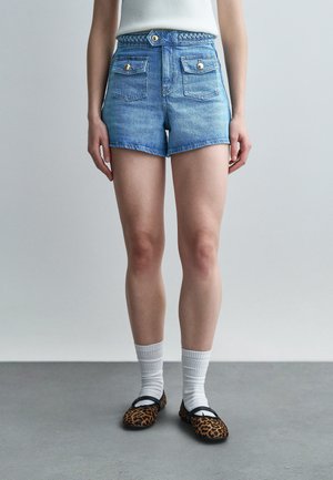 Oseba, oblečena v visoke modre denim kratke hlače z žepi spredaj, bele rebraste nogavice in baletke z leopardjim vzorcem na sivi tla.