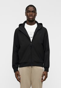 Μαύρο μπουφάν με φερμουάρ και κουκούλα, κατασκευασμένο από απαλό ύφασμα. Διαθέτει ριμπ τελείωμα και μανσέτες, με ένα διακριτικό λογό τυπο στο στήθος. Το μοντέλο στέκεται μπροστά σε απλό φόντο.