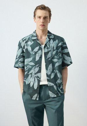 Joven con camisa de manga corta verde oscuro con un ligero estampado de hojas sobre una camiseta blanca sin mangas y pantalones verdes a juego, con las manos en los bolsillos.