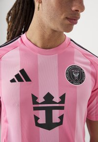 Rosa Sporttrikot mit schwarzen Akzenten, vertikalen Streifen und einem großen schwarzen grafischen Emblem, das einer Krone und einem Anker ähnelt.
