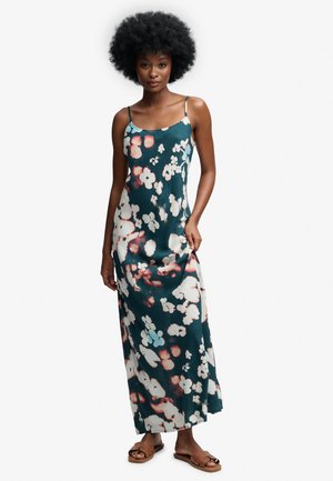 Kobieta z afro w długiej turkusowej sukience na ramiączkach z białymi i różowymi kwiatowymi wzorami oraz brązowych płaskich sandałach na białym tle.
