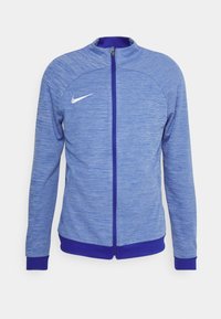 Blå atletisk jakke med lynlås foran, med tekstureret stof, ribstrikkede manchetter og en hvid Nike-logo på venstre bryst.