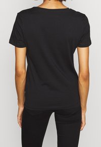 En person iført en ensfarvet sort t-shirt med korte ærmer og sorte bukser, set bagfra mod en ensfarvet lys baggrund.