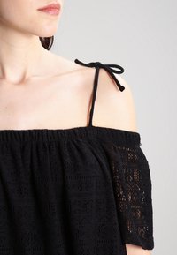 Žena nosí černý top s odhalenými rameny, který má krajkový vzor a tenké ramínko uvázané do mašle na pravém rameni.