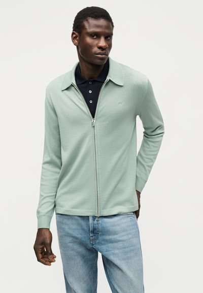 Άνδρας που φοράει ανοιχτοπράσινο πουλόβερ με φερμουάρ πάνω από μαύρο πόλο και ανοιχτό μπλε τζιν, στέκεται μπροστά σε απλό φωτεινό φόντο.