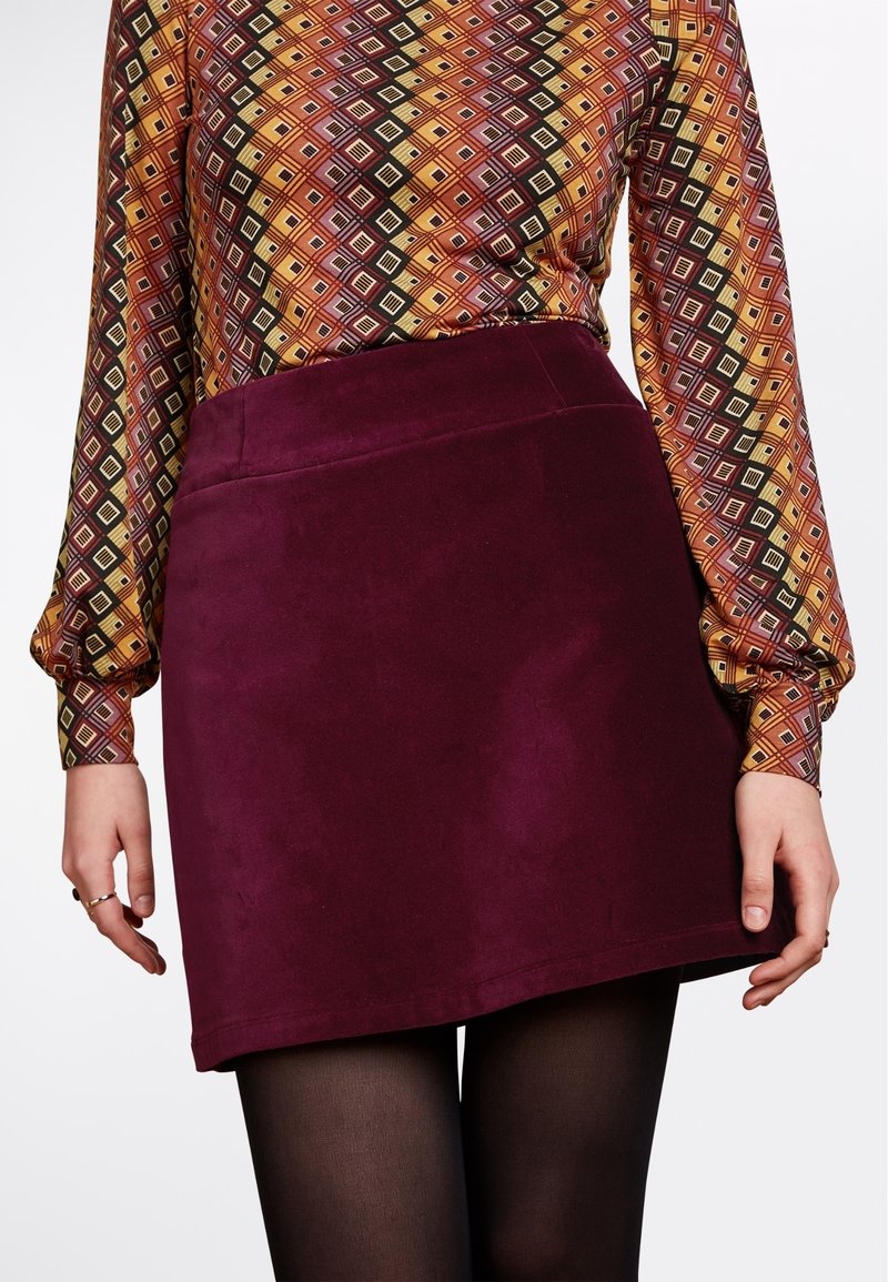Femme portant un haut à manches longues à motifs rentré dans une jupe violet foncé avec des collants noirs, mains détendues sur les côtés.