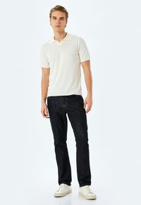 Kortærmet polo shirt i off-white strik, med flad krave, båret sammen med mørk denim jeans med diskrete stikninger og hvide sneakers.