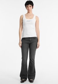 Witte tanktop met een ronde hals en zwarte logo tekst; gecombineerd met donkergrijze uitlopende jeans en zwarte schoenen. Eenvoudig, aangesloten ontwerp.