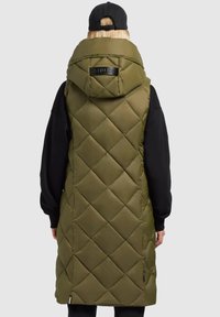 Olivenfarbener gesteppter Padded-Weste mit großer Kapuze, präsentiert mit einem schwarzen Logo-Detail auf der Rückseite und seitlichen Taschen. Getragen über einem schwarzen Langarmshirt.