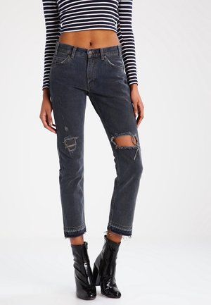 Frau trägt schwarze zerrissene Jeans mit ausgefransten Säumen, gestreiftes, langärmeliges Crop-Top und glänzende schwarze Stiefeletten, steht vor einem einfarbigen Hintergrund.