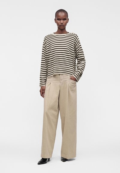 Femme portant un haut à manches longues beige et noir rayé, un pantalon large beige et des chaussures noires pointues, debout avec une main dans la poche.