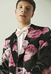 Μαύρο φλοράλ σακάκι με μεγάλα σχέδια ροζ τριαντάφυλλων, συνδυασμένο με λευκό πουκάμισο και ελαφρύ ροζ παπιγιόν, που αναδεικνύει υφασμάτινη υφή και κομμένο μέγεθος.