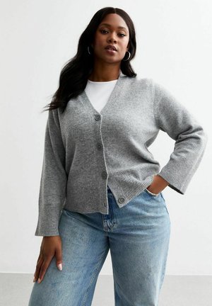 Cardigan grigio lavorato a maglia con scollatura a V, chiusura con bottoni e polsini a coste. Abbinato a jeans azzurri chiari, presenta una vestibilità rilassata e una texture morbida.