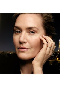 Gros plan sur le visage d'une femme avec une peau lisse et un maquillage léger. Elle a les cheveux châtains clairs et porte une bague en or avec de petits accents.