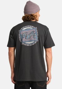Svart bomull T-skjorte med bakre grafikk som viser abstrakte bølger og geometriske mønstre i blått og grått. Modellen har på seg en lavendel lue.
