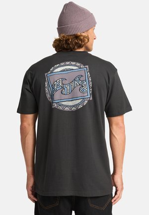 Zwarte katoenen t-shirt met een achterafbeelding van abstracte golven en geometrische patronen in blauw en grijs. Model draagt een lavendel-beanie.