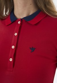 Punane polo särk, millel on tekstuuriga kangas, tumesinine krae, neli nuppe ja vasakul rinnal tume sinine linnulogo broderitud.