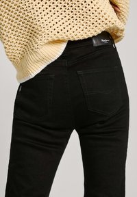 Jeans negros con un diseño ajustado, que cuentan con dos bolsillos traseros, costuras sutiles y un parche de cuero con el logo en la cintura.