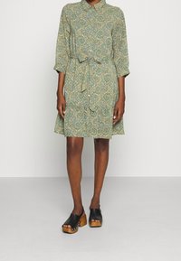 Vihreä kukallinen mekko, jossa on kauluspaita, vyötärönauha ja kolmannekselle asti ulottuvat hihat. Yhdistetty mustiin korokekengiin ja puupritsikengiin.