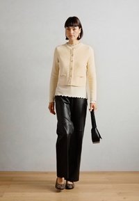 Beige strikket cardigan med knapper, sammen med svarte skinnbukser og dyre-print leiligheter, holder en svart håndveske. Minimalistisk bakgrunn.