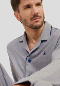 Parte superior de pijama a rayas grises con ribete azul, con un frente de botones y detalle de logo. Textura suave, diseño de cuello clásico, corte relajado.