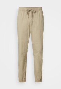 Beige lichtgewicht broeken met een elastische tailleband en aantrekkoord, met taps toelopende benen en elastische boorden bij de enkels. Gladde textuur.