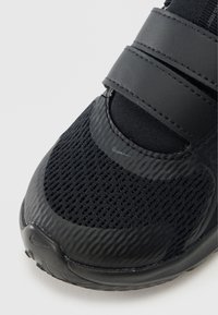 Zapato deportivo negro con malla y material texturizado, cuenta con dos correas de Velcro y una suela de goma lisa. Diseño de punta redonda.
