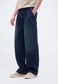 Άτομο που φοράει σκούρο μπλε τζιν παντελόνι με φαρδιά μπατζάκια, μπεζ αθλητικά παπούτσια και κρεμ πουκάμισο, με τα χέρια στις τσέπες, στέκεται μπροστά σε μονόχρωμο φόντο.