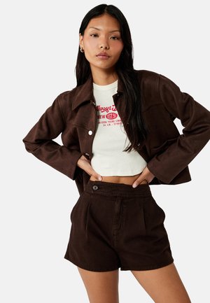 Ung kvinde iført en cropped hvid grafisk T-shirt, mørkebrun jakke med knapper og højtaljede mørkebrune shorts, poserer med hænderne på hoften.