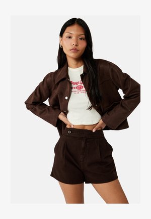 Jeune femme portant un t-shirt graphique blanc court, une veste marron foncé boutonnée et un short marron foncé taille haute, posant les mains sur les hanches.