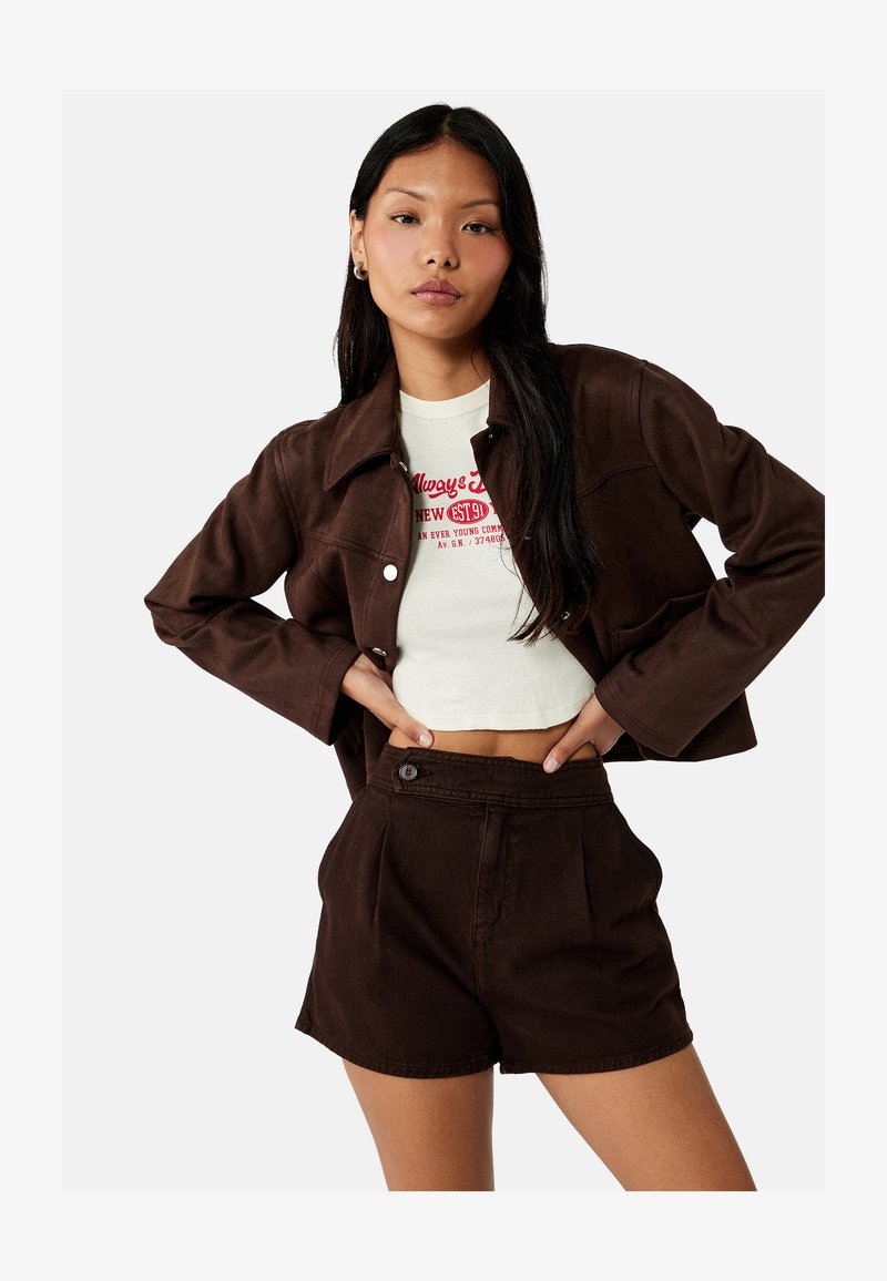 Young woman wearing a cropped white graphic T-shirt, dark brown buttoned jacket, and high-waisted dark brown shorts, posing with hands on hips.