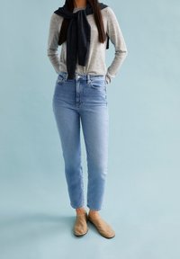 Chemise à manches longues gris clair avec un pull gris foncé noué au cou, jean taille haute bleu clair et chaussures slip-on beige.