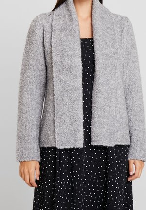 Cardigan grigio a maglia con una texture morbida e soffice, apertura frontale, maniche lunghe e senza chiusure, indossato sopra un vestito nero a pois.