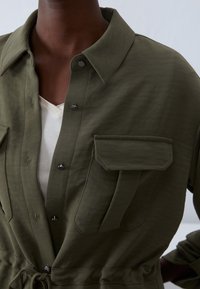Olive green shirt jacket made of lightweight fabric, featuring a pointed collar, snap buttons, and two large chest pockets with a belted waist.