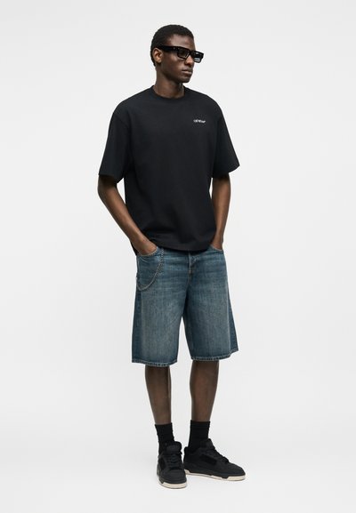 Άνδρας που φοράει μαύρα γυαλιά ηλίου, μαύρο φαρδύ μπλουζάκι, τζιν σορτς μέχρι το γόνατο με αλυσίδα, μαύρες κάλτσες και μαύρα αθλητικά παπούτσια, στέκεται ακίνητος.