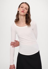 Hvid langærmet top med bred scoop-udskæring, lavet af et blødt, glat stof. Kantene er buede og den er parret med en sort nederdel.