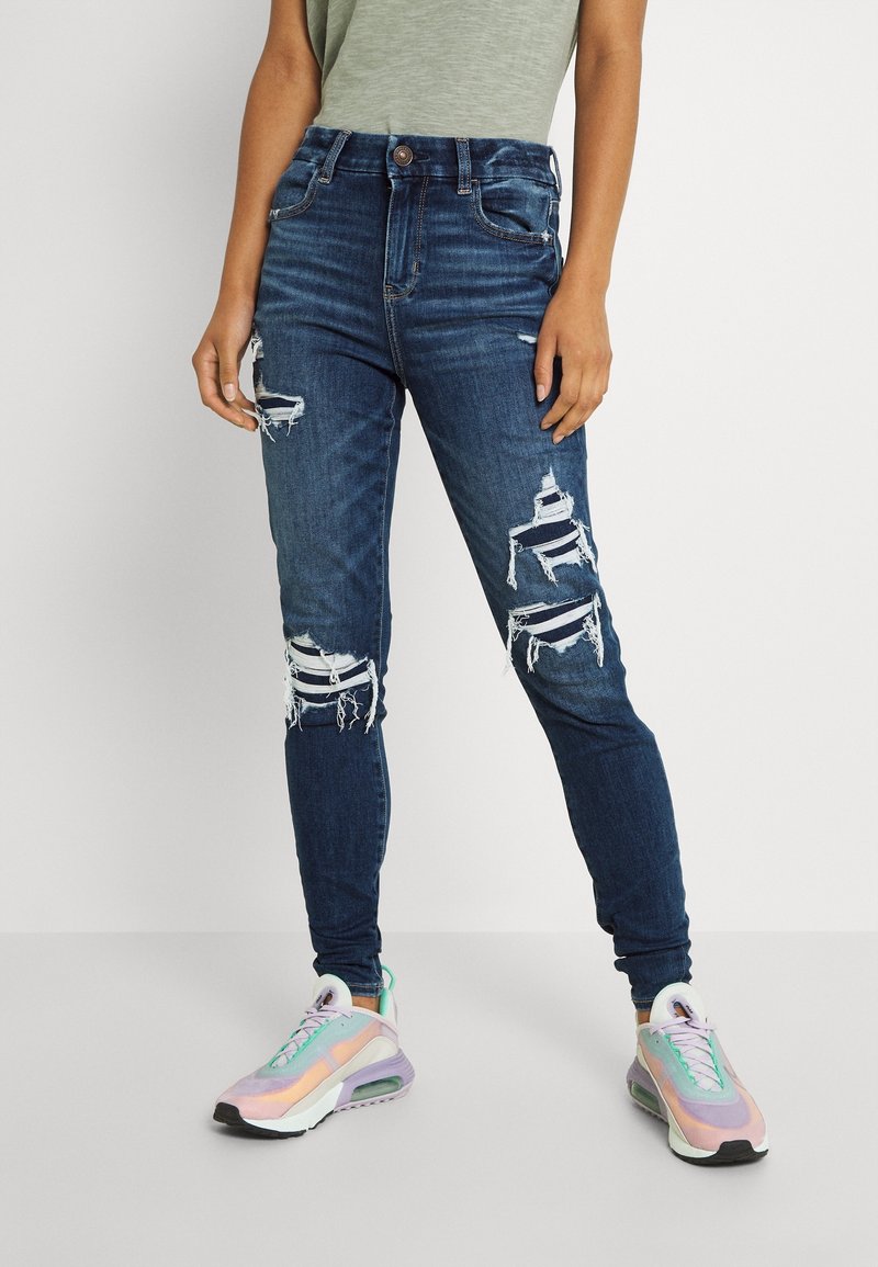 Personne portant un jean skinny bleu foncé déchiré et des baskets multicolores, debout sur un fond blanc.