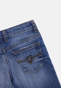 Lato posteriore di jeans blu in denim con cuciture marroni e una piccola toppa triangolare con il logo GUESS sulla tasca.